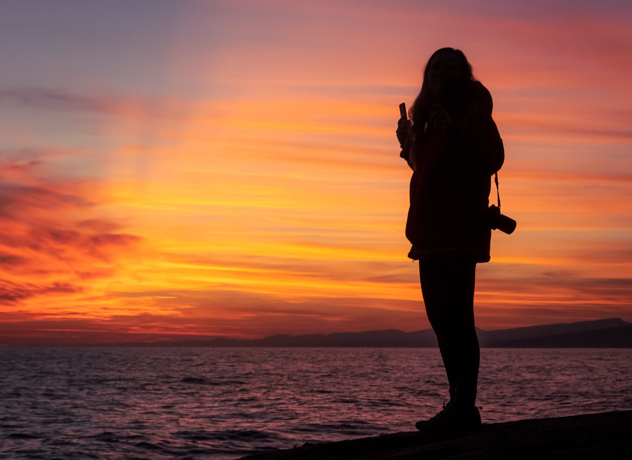 Where to watch the sunset in Grand Marais ? Head out to artist point. Image of a photographer watching a beautiful red sunset over Lake Superior with the sawtooth mountains outlined in the background