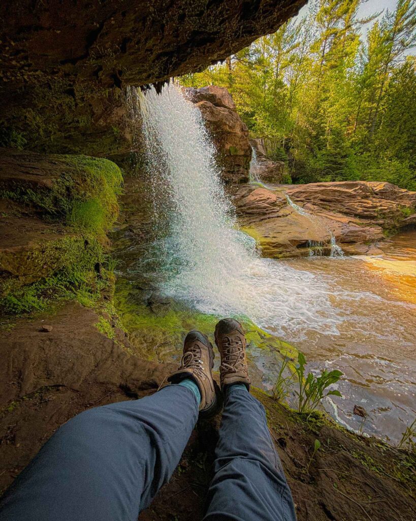 image of hiker's legs and boots facing the cave under o kun de kun falls in the keweenaw peninsula