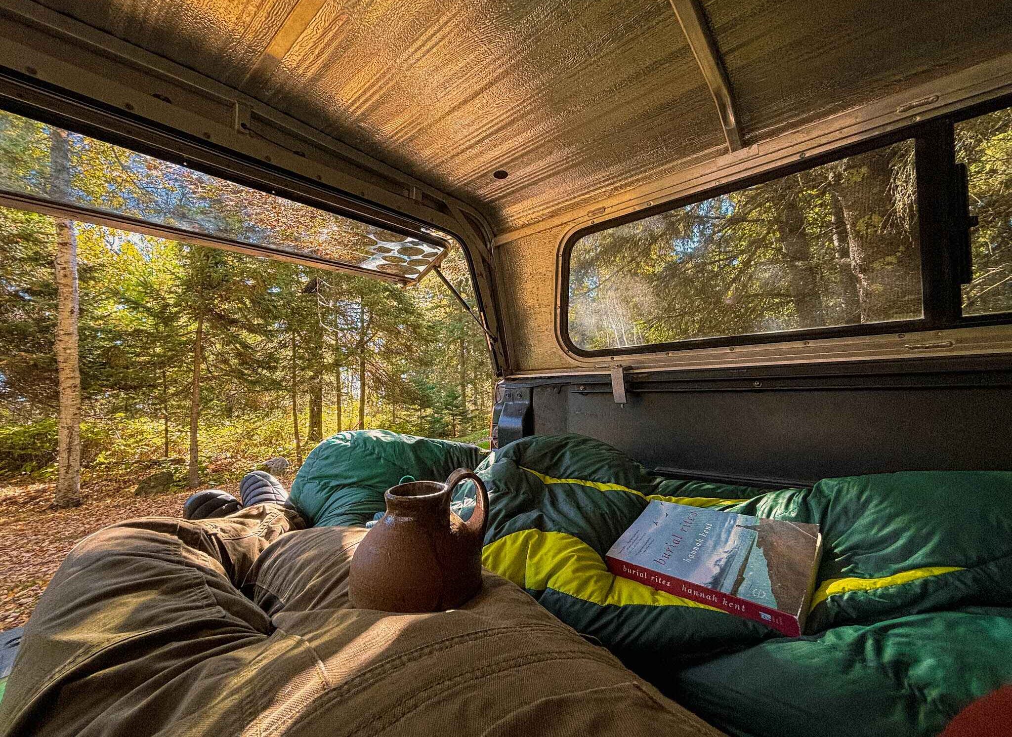 image of a car camping travel set up in the late autumn woods. A mug sits on a womans lap, a sleeping bag curled at her side with a book on top.
