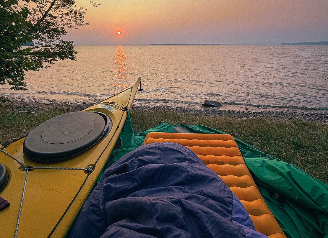image of a cowboy camp at sunrise. A sleeping bag sits on top of a sleeping pad next to a sea kayak in the Apostle Islands